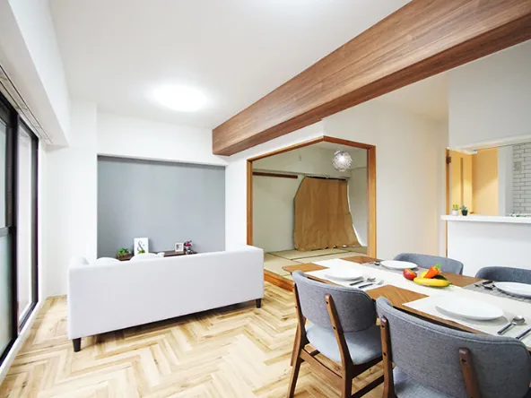 写真：4人掛けのダイニングテーブルと壁際に白いテーブルがあるフローリングの部屋。奥には一段上がった畳み敷きの部屋がある